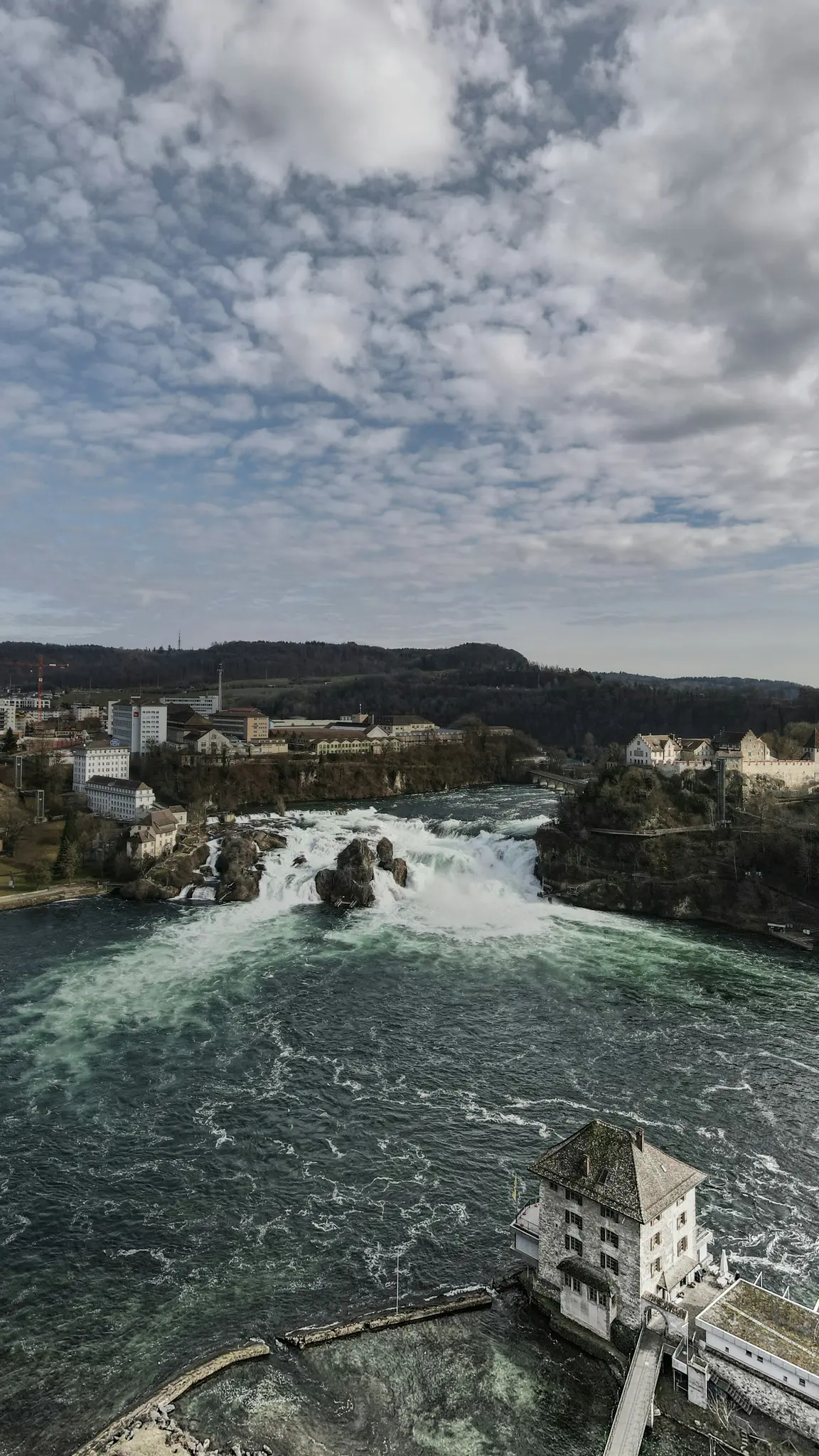 An aerial shot shows a powerful waterfall cascading over rocks in a wide river, flanked by buildings and forested hills.
