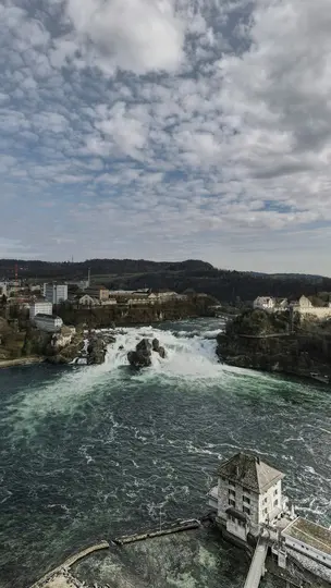 An aerial shot shows a powerful waterfall cascading over rocks in a wide river, flanked by buildings and forested hills.