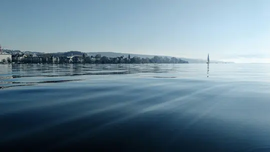 Široka pejzažna fotografija mirnog, plavog jezera s blagim valovima. Na udaljenoj obali vidi se gradski obris i brda…