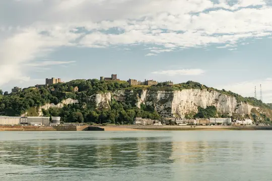 Ett brett landskapsfoto visar en kuststad med vita klippor, ett slott på en grön kulle och byggnader längs stranden.