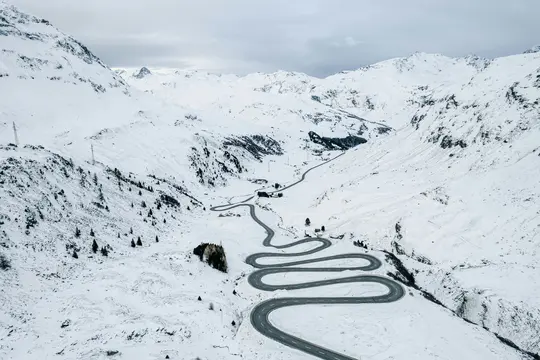 Una vista aérea muestra una carretera oscura y sinuosa que serpentea a través de un vasto paisaje montañoso cubierto de nieve