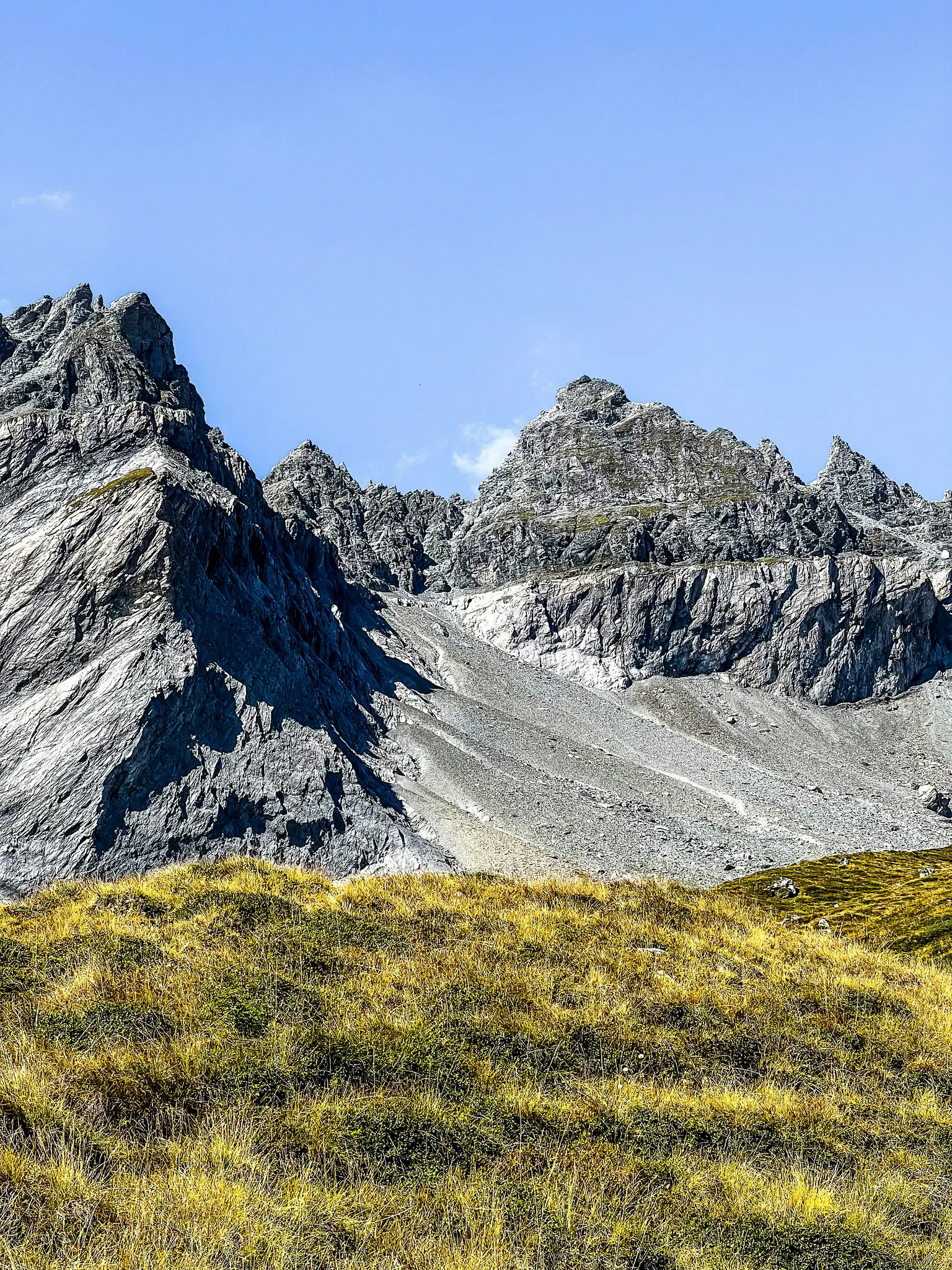 A wide landscape view of jagged, grey rocky mountains under a clear blue sky, with sunlit golden and green grass in the…