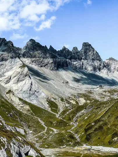 A wide landscape view of a mountain range with sharp, grey rocky peaks under a bright blue sky with scattered white clouds.…