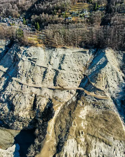 An aerial shot captures a rugged, eroded hillside with a winding path, leading up to a village nestled among trees.