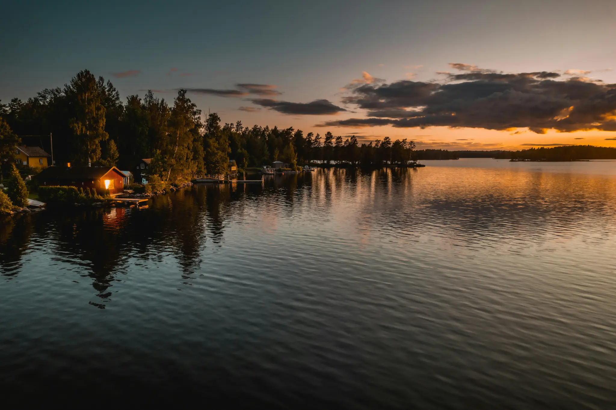 A wide landscape photo of a calm lake at dusk. A red house with a glowing window sits on the dark, tree-lined shore.