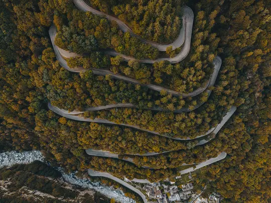 An aerial shot shows a serpentine road cutting through a dense autumn forest, leading to a small village by a rocky river.