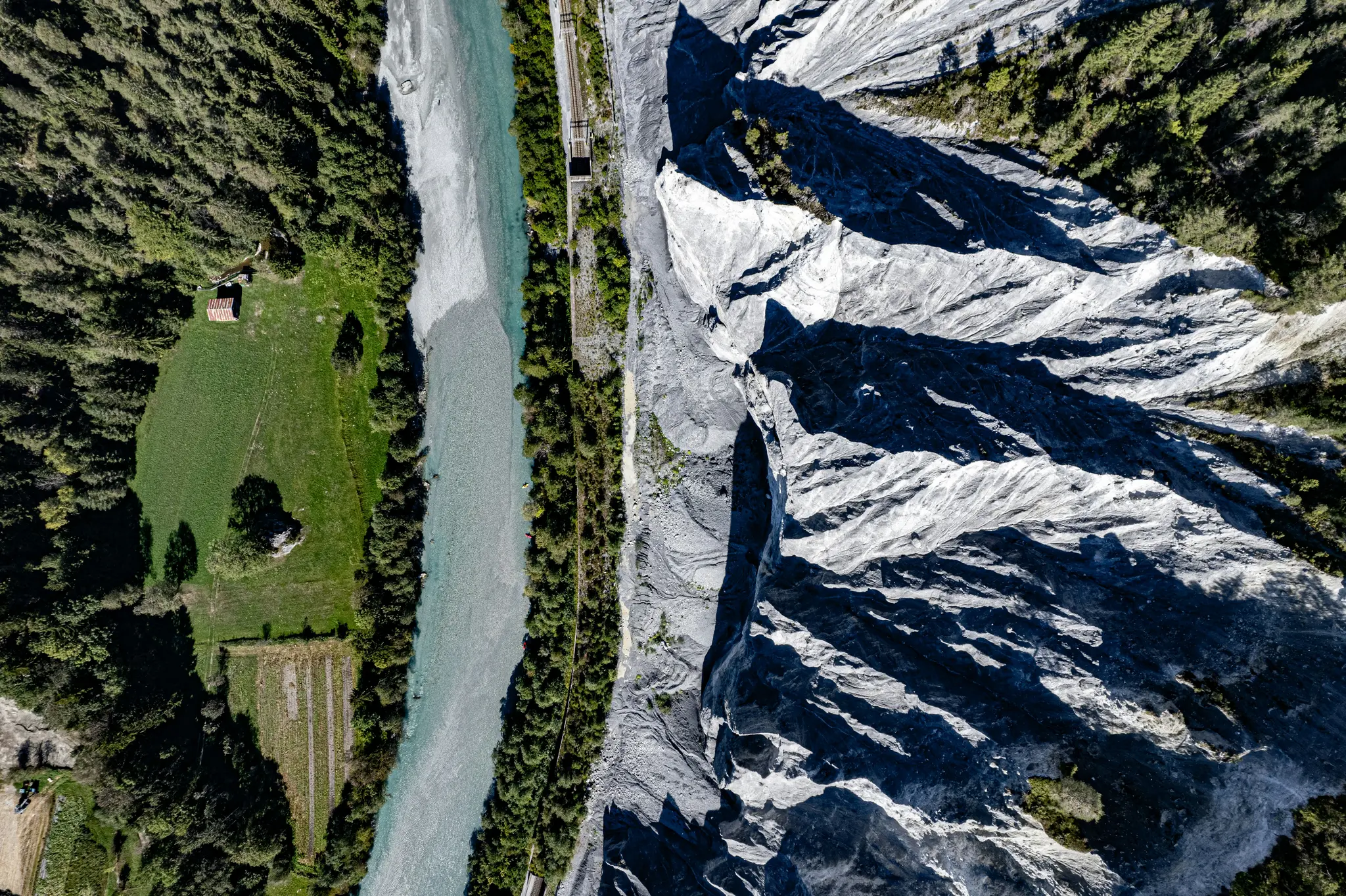 Ett flygfoto visar en ljusblå flod som skiljer en tät grön skog och en grå, tandad klippvägg. Ett litet rött tak...