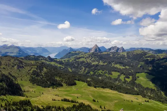 An aerial landscape photo shows rolling green hills covered with dark green trees, leading to jagged mountains and a blue…