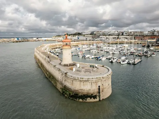 Una vista aérea muestra un muelle de piedra curvo con un pequeño faro en su extremo, que encierra un puerto deportivo lleno