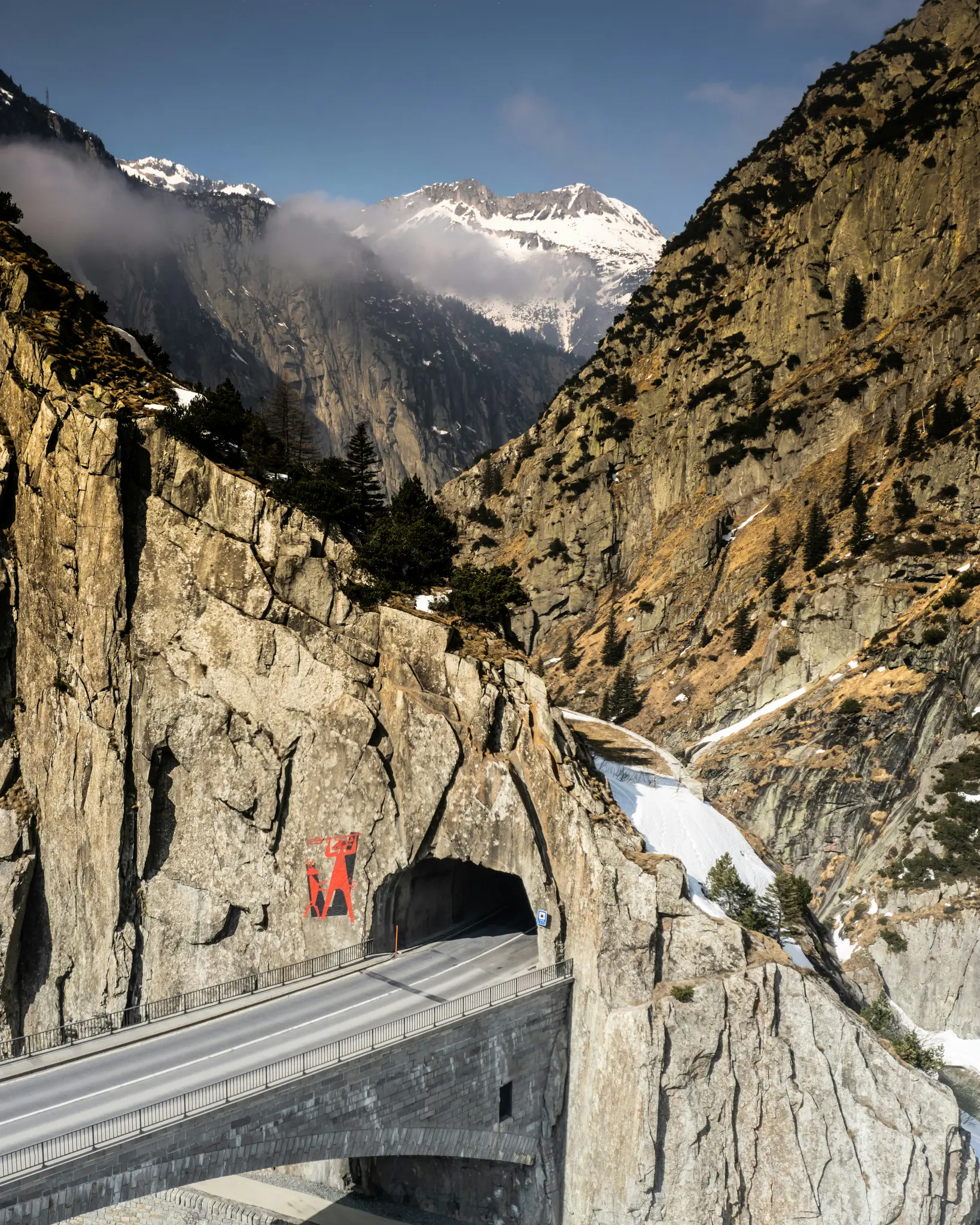 En slingrande bergsväg leder in i en mörk tunnel huggen i en massiv klippvägg, flankerad av branta, snötäckta berg.