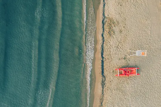 Una toma aérea muestra una playa de arena con suaves olas a la izquierda, un bote de rescate rojo y una silla de socorrista