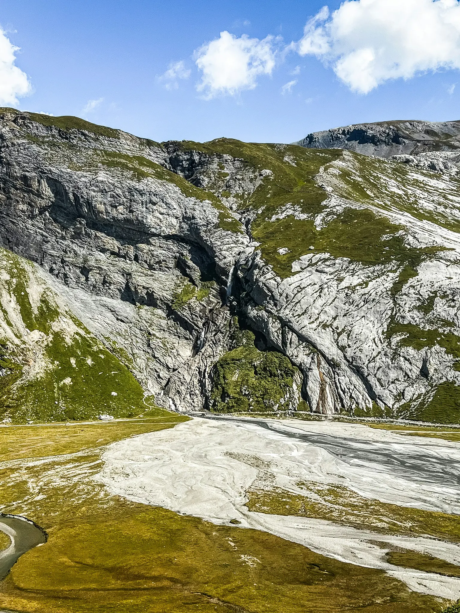 A wide landscape photo shows a dry, light-colored riverbed winding through green grassy slopes at the base of rocky…