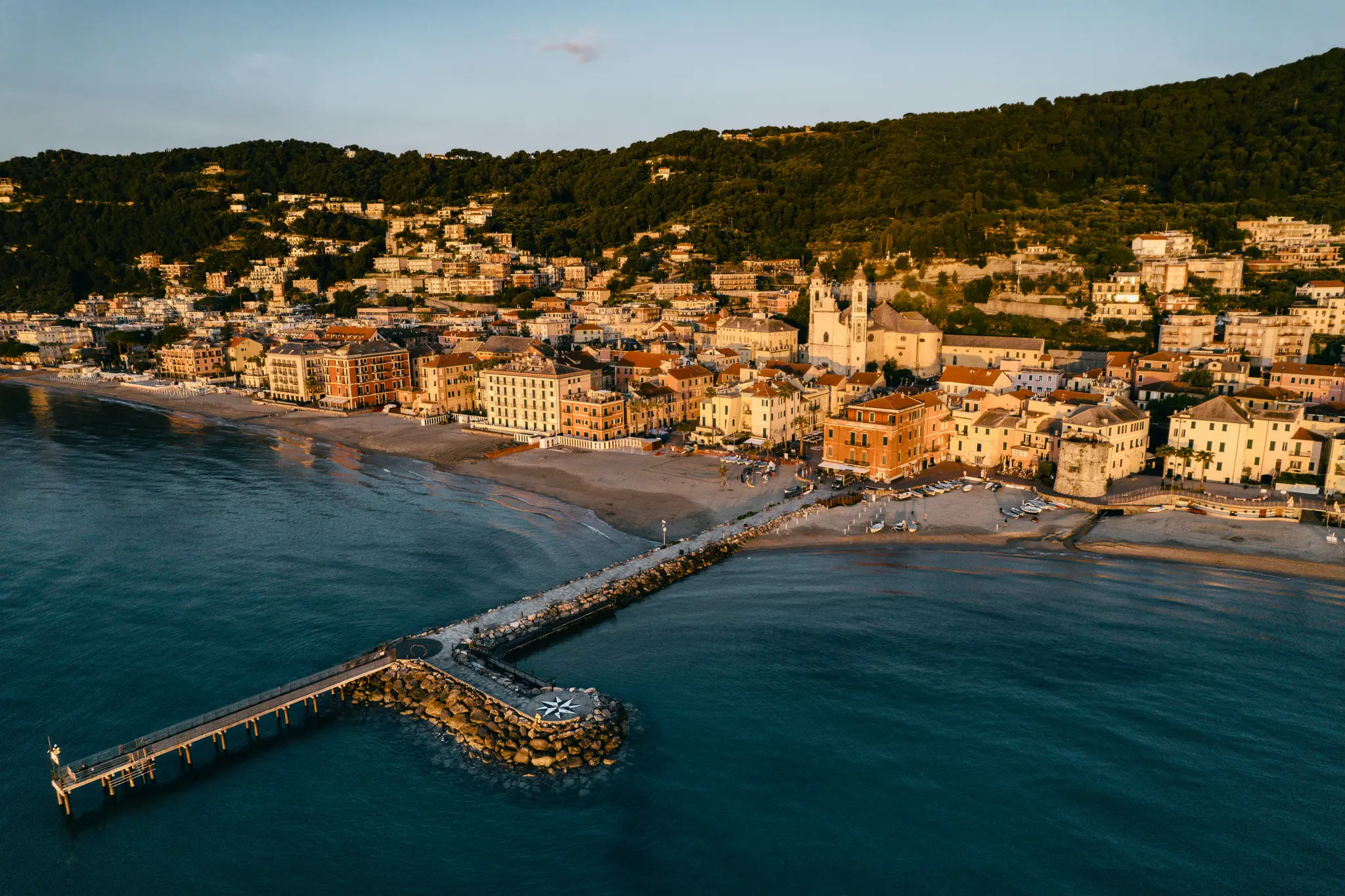 An aerial shot of a vibrant coastal town with colorful buildings nestled against a green hill, a long pier stretches into…