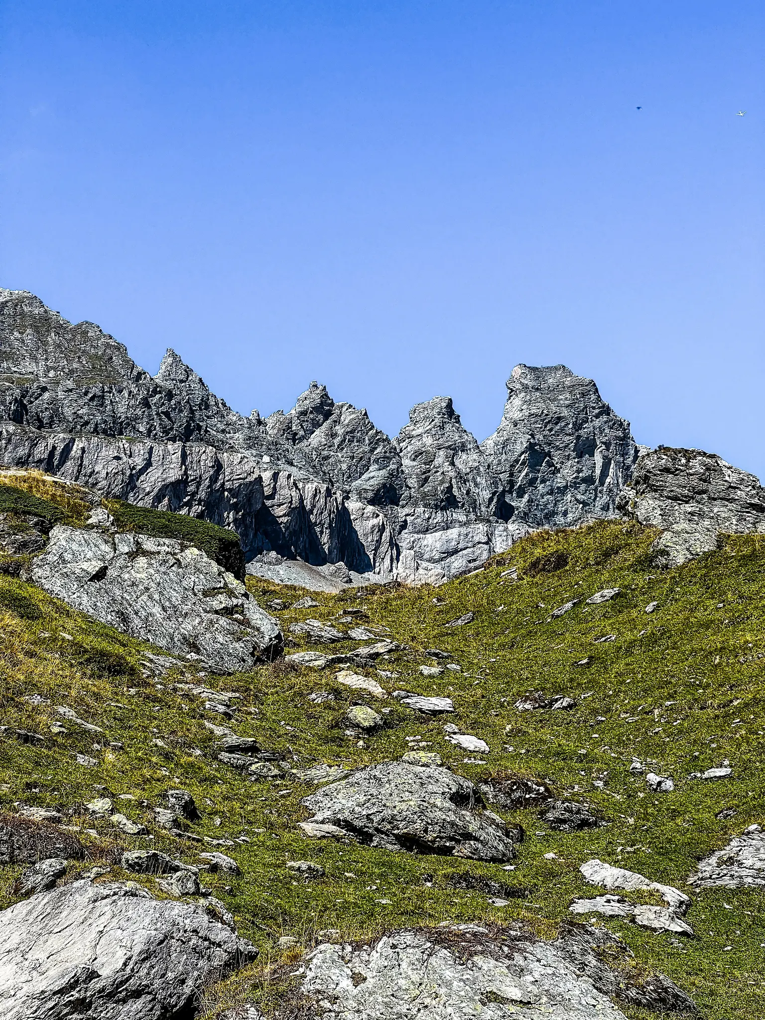 A vibrant photo of a mountain landscape with a steep, rocky, green slope leading up to jagged grey peaks under a clear blue…