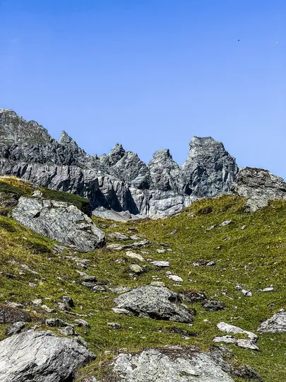 A vibrant photo of a mountain landscape with a steep, rocky, green slope leading up to jagged grey peaks under a clear blue…