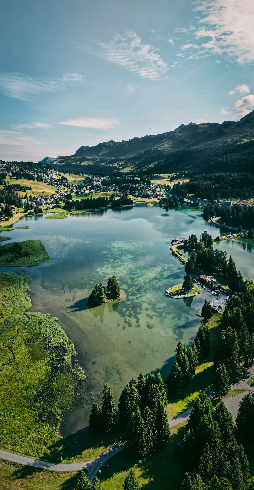 An aerial shot captures a clear, turquoise lake nestled among lush green mountains and a small village under a blue sky.
