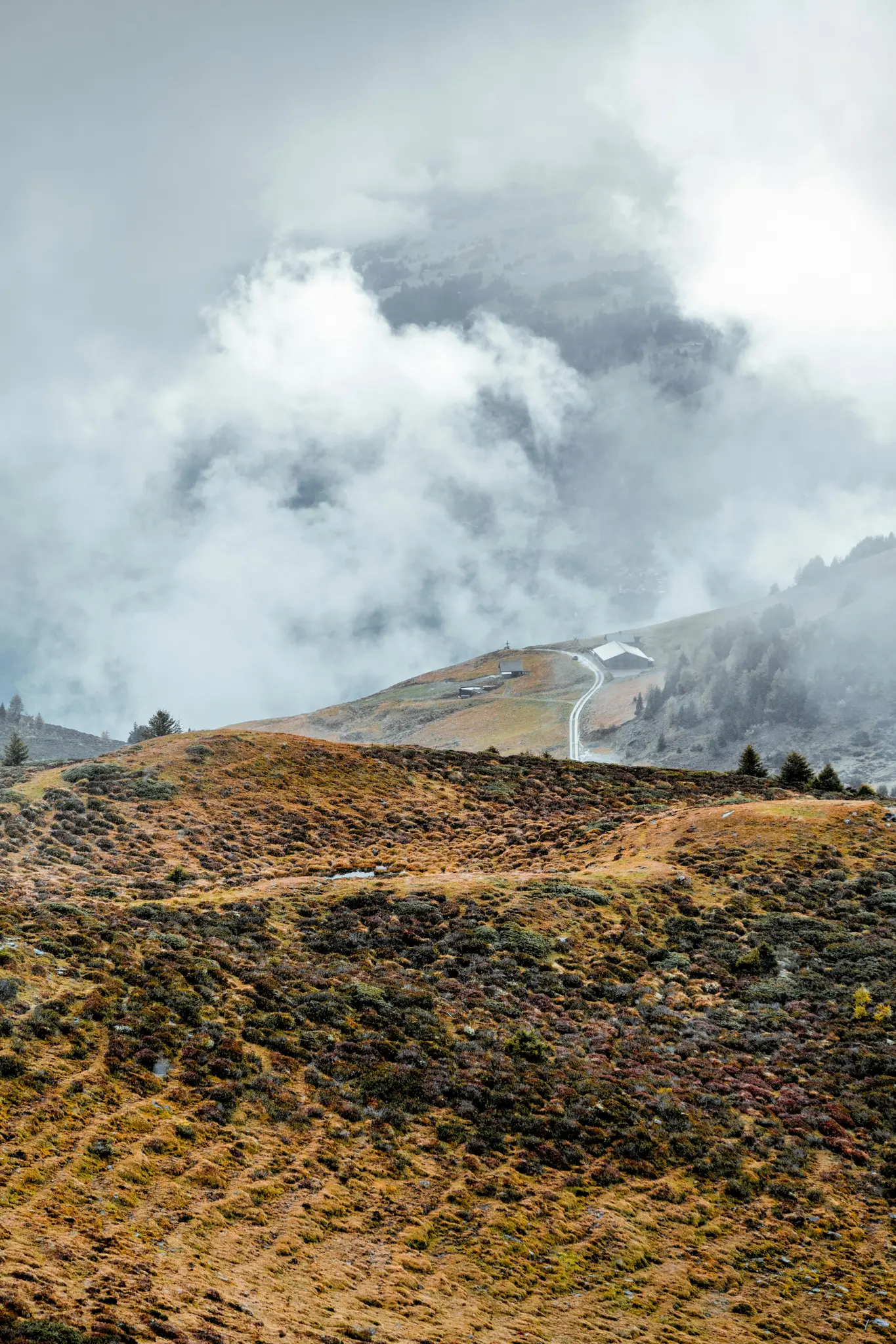 A winding road ascends a mountain slope covered in orange and green shrubs, leading to buildings under a cloudy sky.