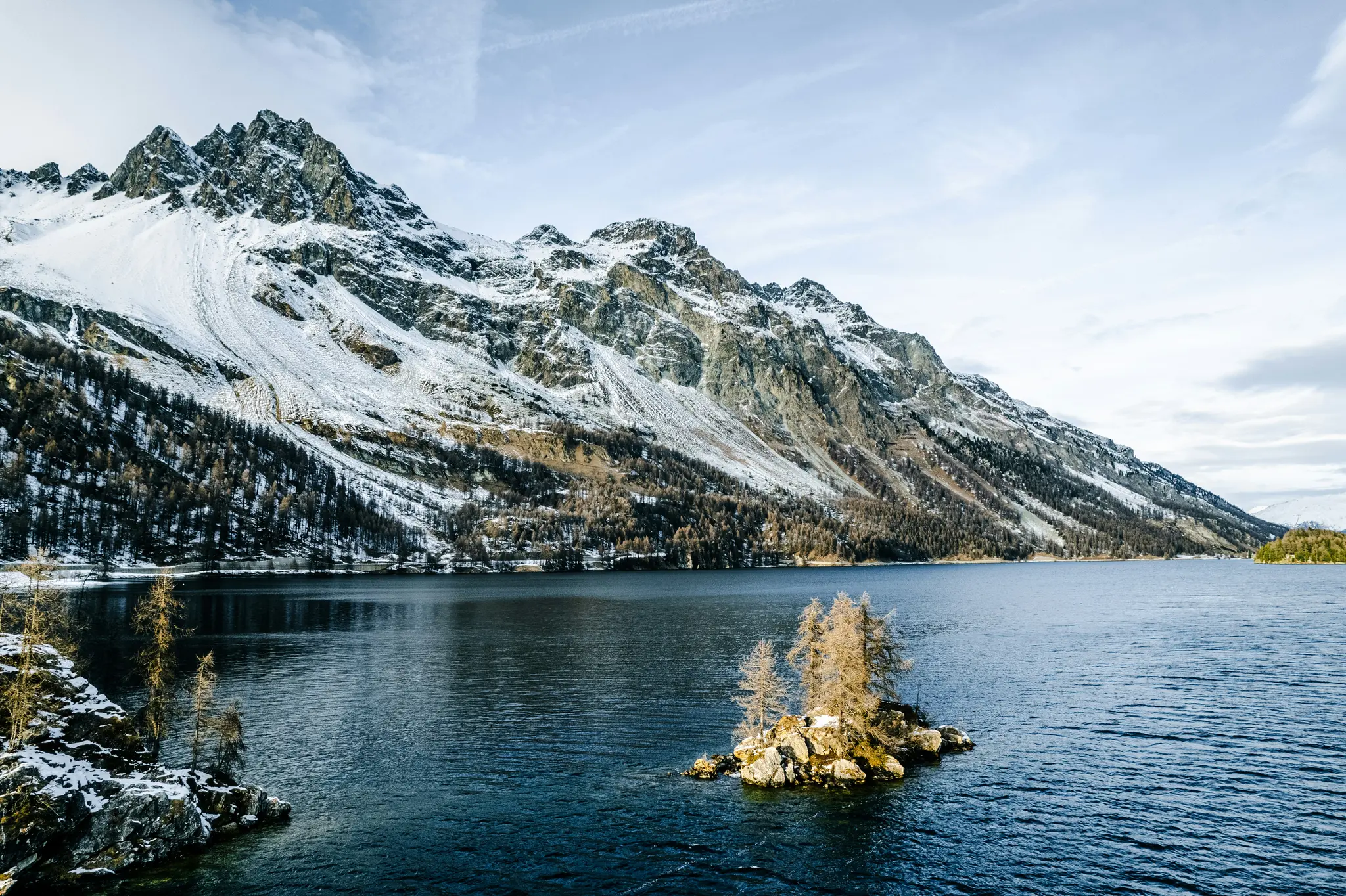 A wide shot of a vast, dark blue lake reflecting a towering, snow-covered mountain range under a cloudy sky. A small rocky…
