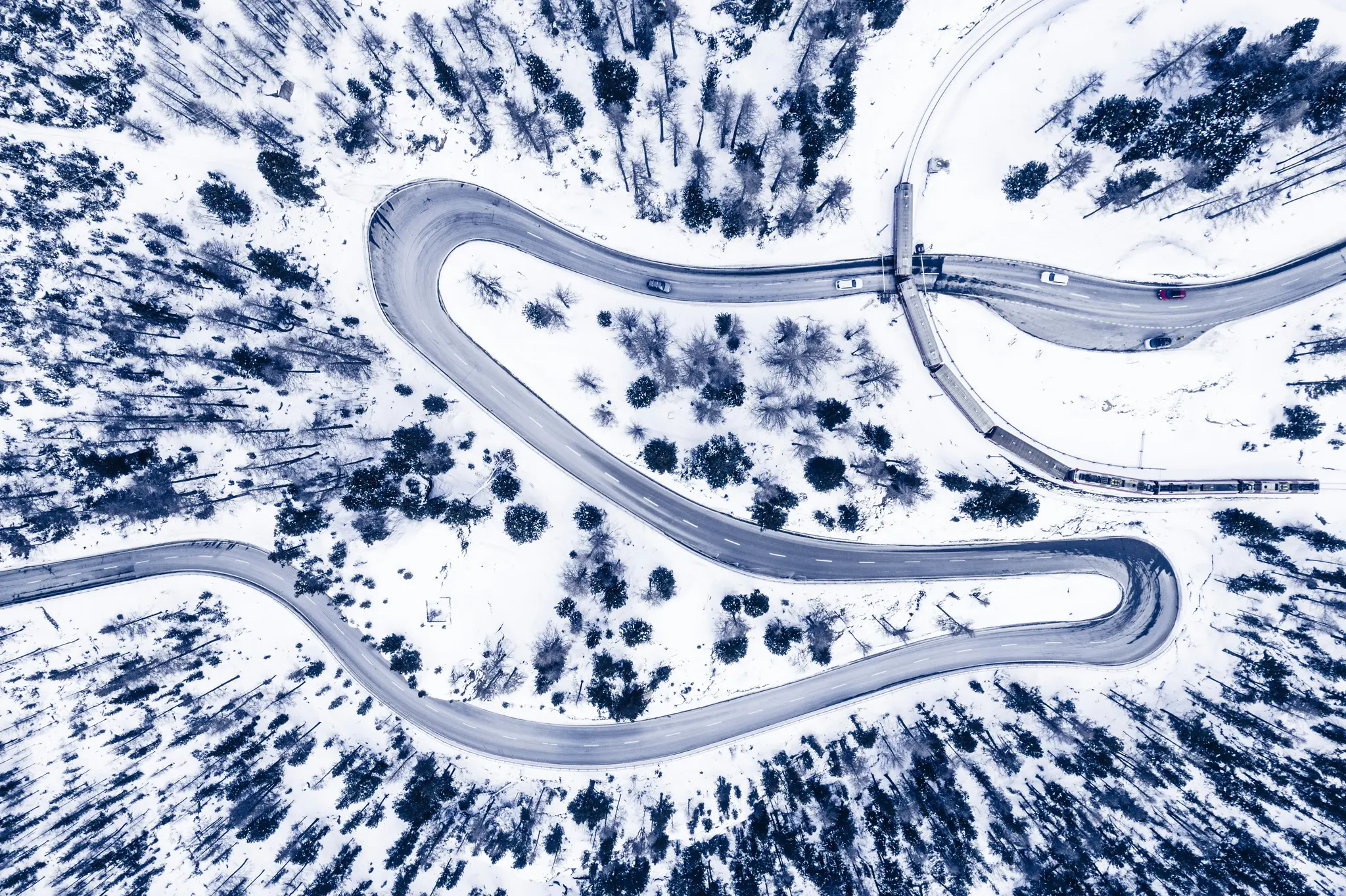 Una vista aérea muestra una carretera sinuosa y una vía de tren que atraviesan un bosque cubierto de nieve. Se ven coches y
