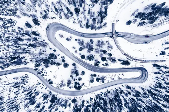 Una vista aérea muestra una carretera sinuosa y una vía de tren que atraviesan un bosque cubierto de nieve. Se ven coches y
