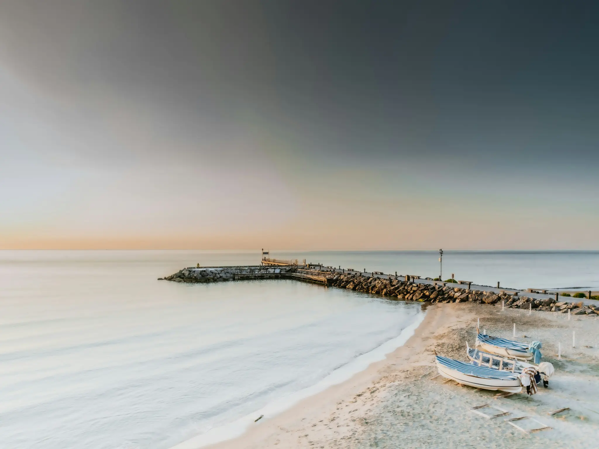 Ett långtidsexponeringsfoto av en lugn strand med en stenpir som sträcker sig ut i havet under en mjuk, gradvis himmel under