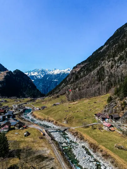 Una vista aérea de un valle verde con un río rocoso que lo atraviesa, un pequeño pueblo y montañas nevadas distantes.