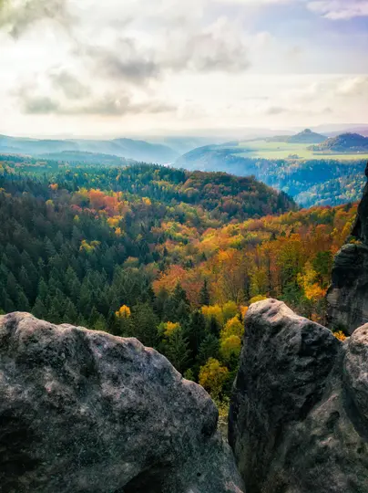 Eine weite Landschaftsaufnahme, die ein Tal voller Herbstbäume in verschiedenen Grün-, Gelb- und Orangetönen zeigt, gesehen