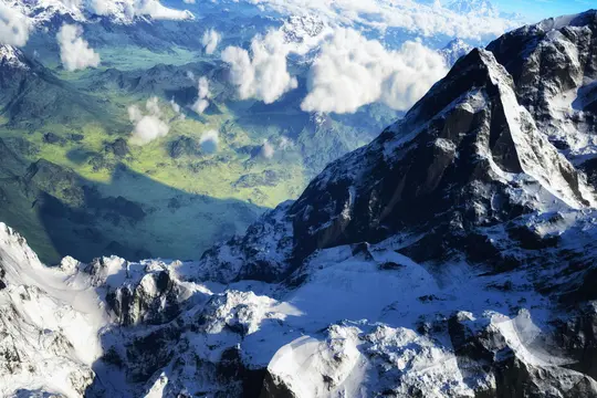 Eine Luftaufnahme zeigt raue, schneebedeckte Berggipfel im Vordergrund, mit einem üppigen grünen Tal, das sich in die Ferne