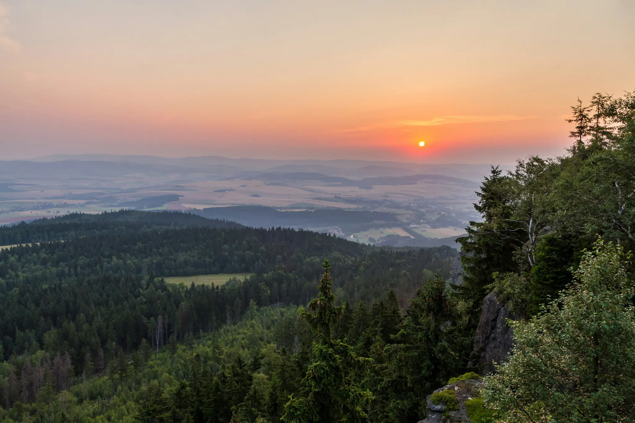 Eine weite Landschaftsaufnahme, die einen lebhaften orange-rosa Sonnenuntergang über einem Tal mit dunkelgrünen Wäldern und