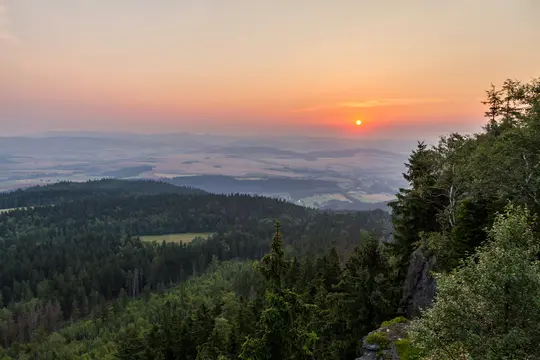 Eine weite Landschaftsaufnahme, die einen lebhaften orange-rosa Sonnenuntergang über einem Tal mit dunkelgrünen Wäldern und
