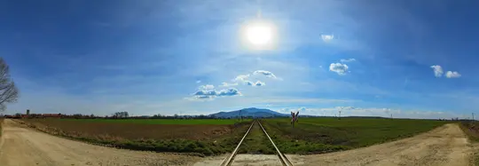 Eine weite Panoramaaufnahme einer ländlichen Szene. Eine einzelne Eisenbahnschiene verläuft durch die Mitte, flankiert von