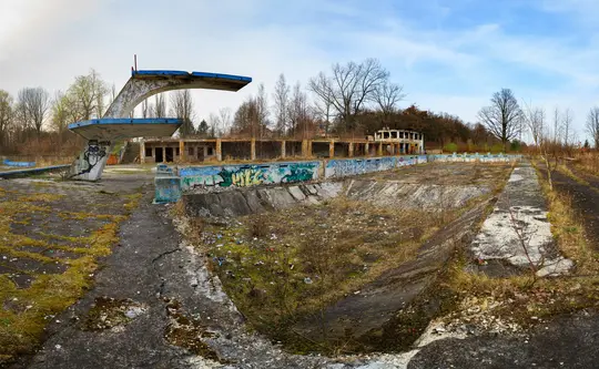 Eine Weitwinkelaufnahme eines verfallenen Freibadkomplexes. Das leere Poolbecken ist mit Unkraut und Schutt überwuchert. Ein