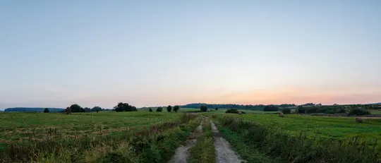 Et bredt landskabsfoto af en grusvej, der skærer gennem levende grønne marker. Himlen ovenover skifter fra lyseblå til…