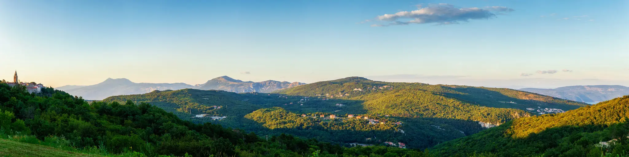 A wide panoramic shot of verdant, rolling hills under a clear sky. A small town with a tall bell tower sits on a hill to…