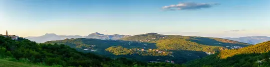 A wide panoramic shot of verdant, rolling hills under a clear sky. A small town with a tall bell tower sits on a hill to…