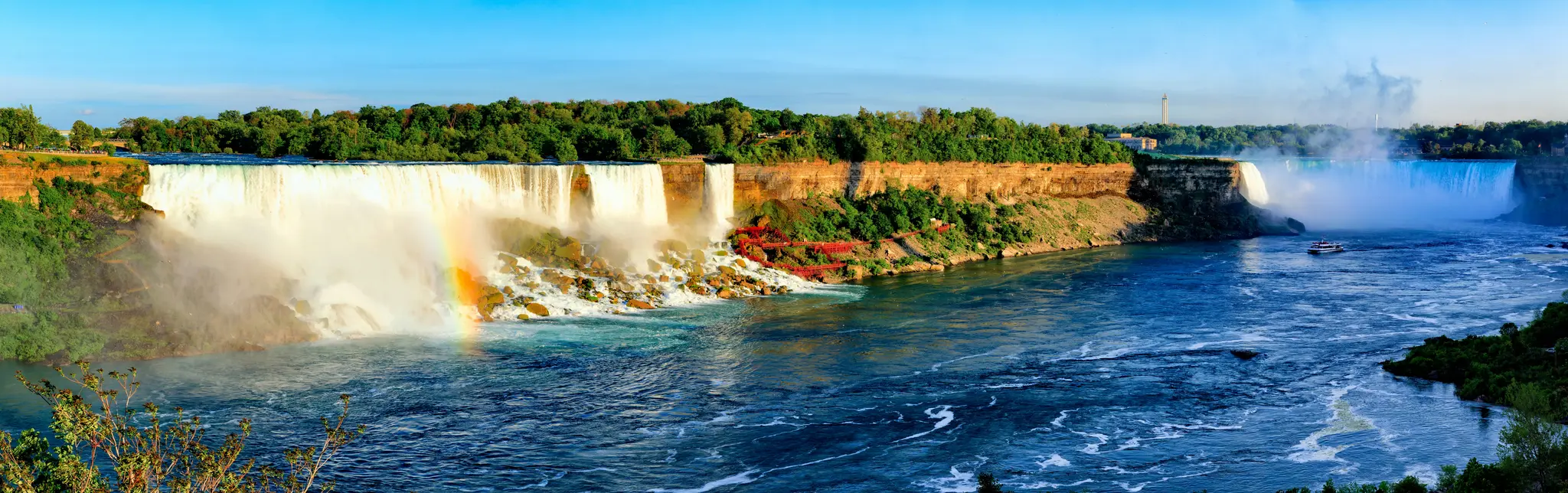 Eine weite Panoramaaufnahme der Niagarafälle unter einem klaren blauen Himmel, mit einem lebendigen Regenbogen, der sich
