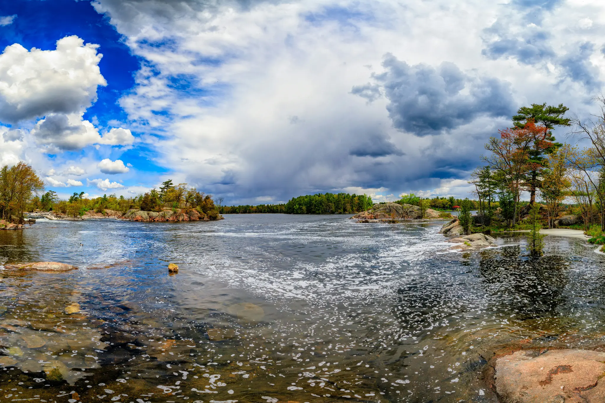 A wide panoramic photo of a river with foamy water flowing around rocky islands and shores, under a sky with both blue…