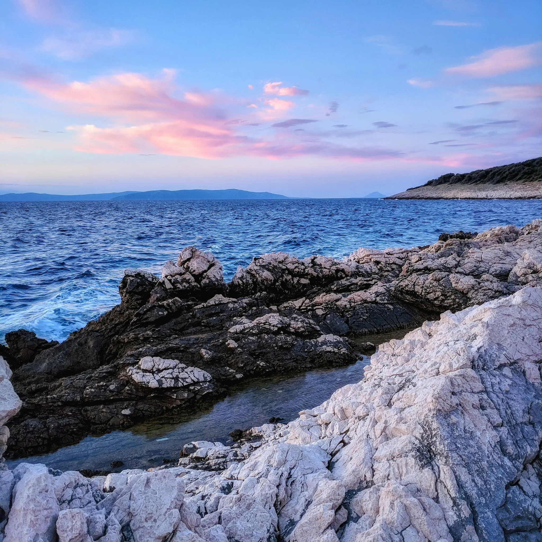 Una vista panorámica de una costa rocosa con agua azul oscuro. El cielo arriba es azul claro con suaves nubes rosadas. Tierra