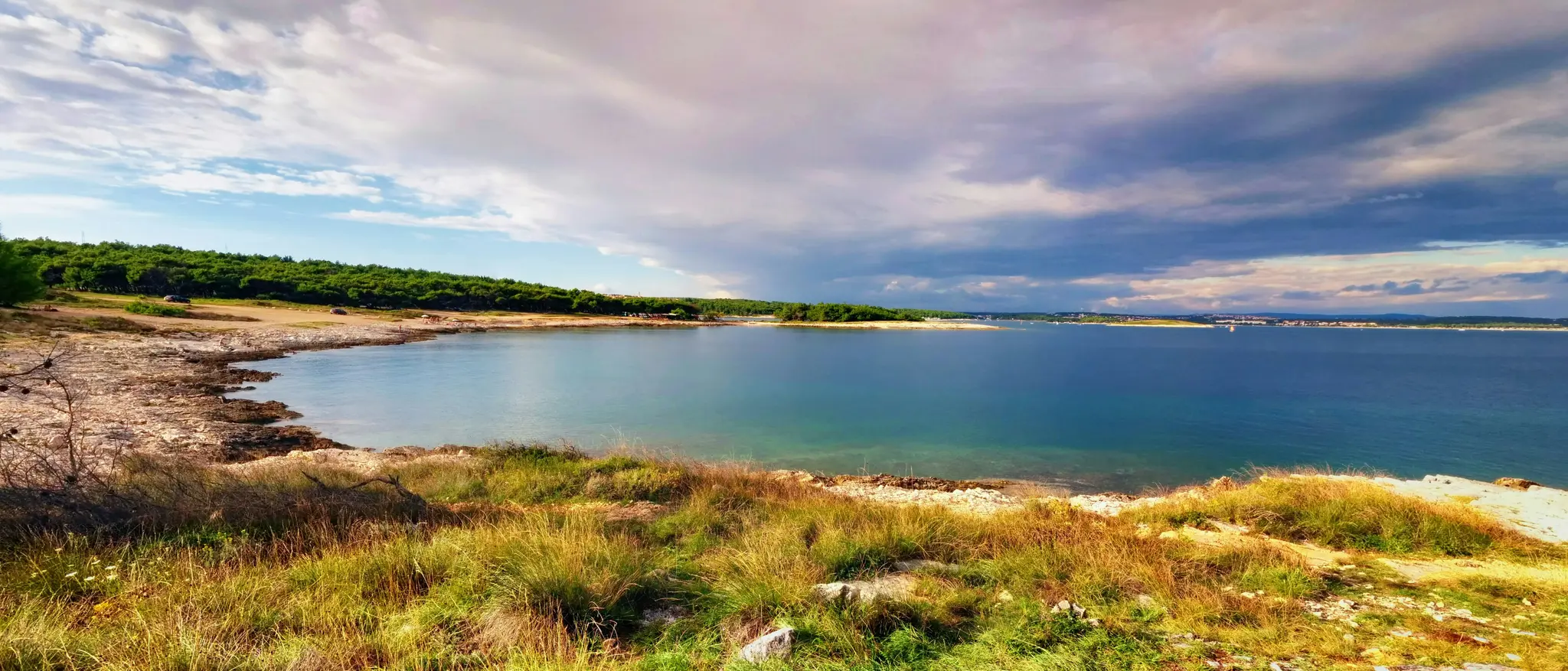 A wide landscape photo of a tranquil bay. The foreground features dry grass and rocks, leading to a rocky shoreline. The…