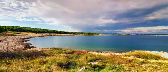 A wide landscape photo of a tranquil bay. The foreground features dry grass and rocks, leading to a rocky shoreline. The…