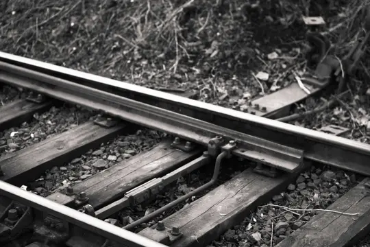Primer plano en blanco y negro de un desvío de vía de tren, mostrando los raíles metálicos, traviesas de madera y balasto de