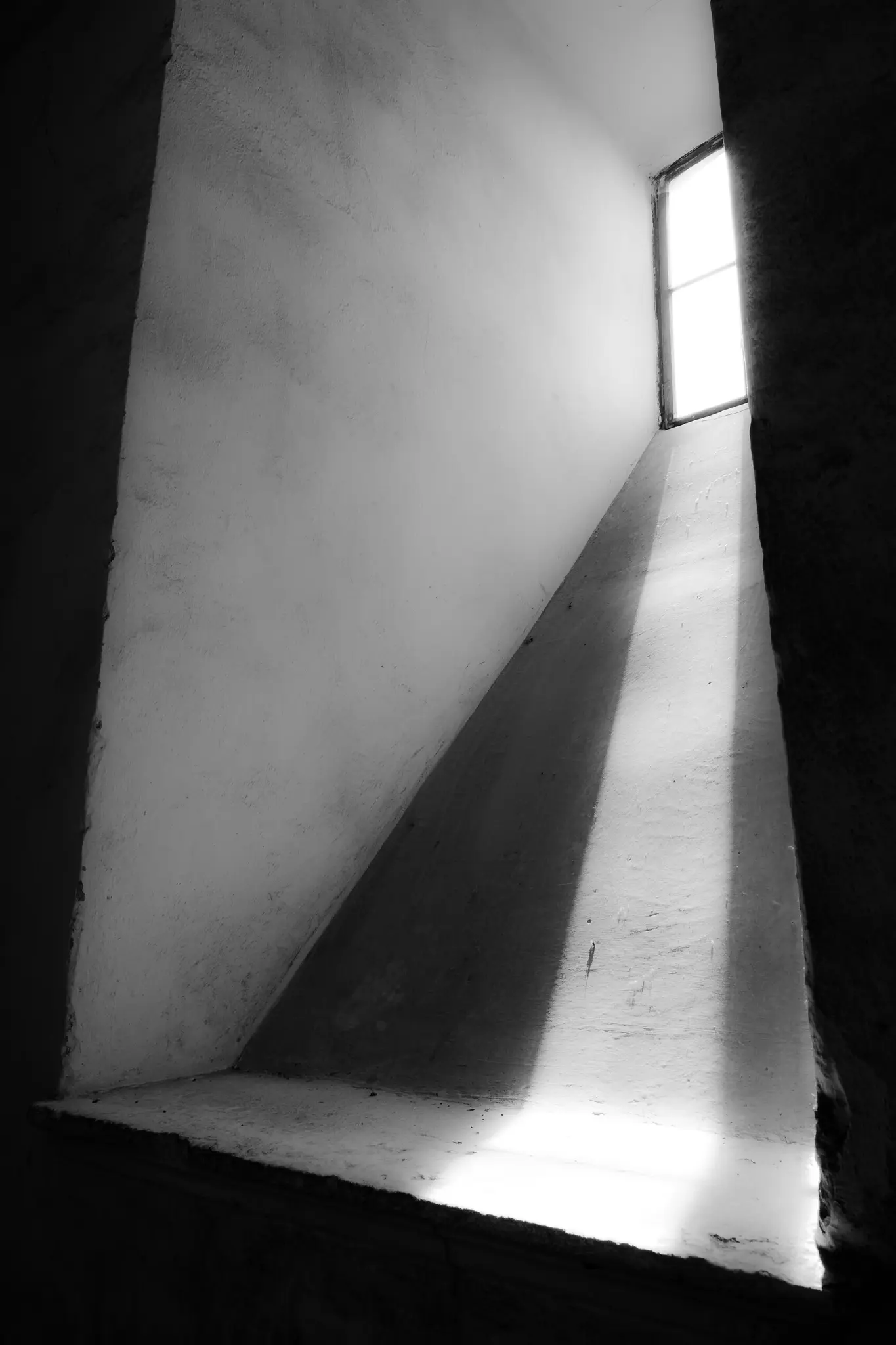 Una foto en blanco y negro muestra una ventana estrecha y alta que proyecta rayos de luz brillantes y angulares sobre una