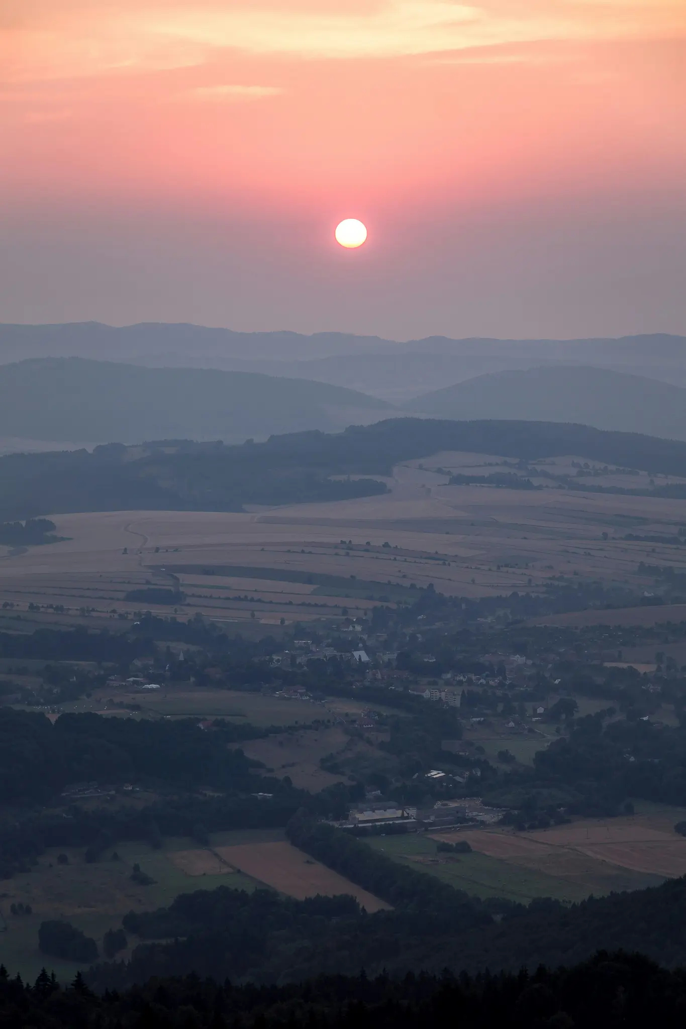 Vista aérea de un paisaje rural al atardecer, con un sol brillante en un cielo rosa y naranja sobre colinas onduladas y...