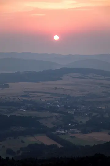 Vista aérea de un paisaje rural al atardecer, con un sol brillante en un cielo rosa y naranja sobre colinas onduladas y...
