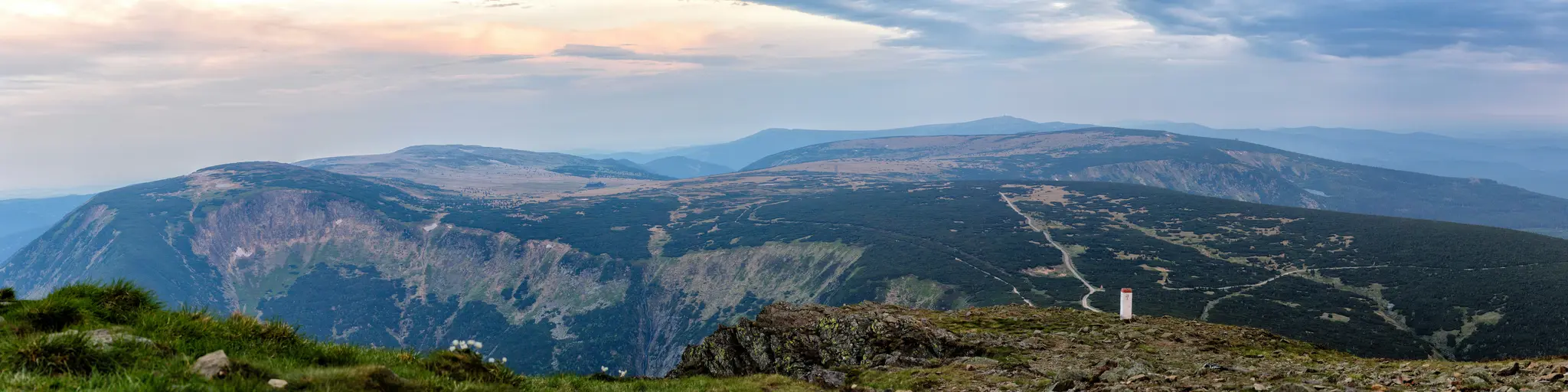 Eine weite Panoramaaufnahme eines Gebirgszugs. Der Vordergrund zeigt felsiges Gelände mit Flecken grünen Grases. Ein tiefes
