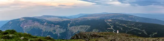 Eine weite Panoramaaufnahme eines Gebirgszugs. Der Vordergrund zeigt felsiges Gelände mit Flecken grünen Grases. Ein tiefes