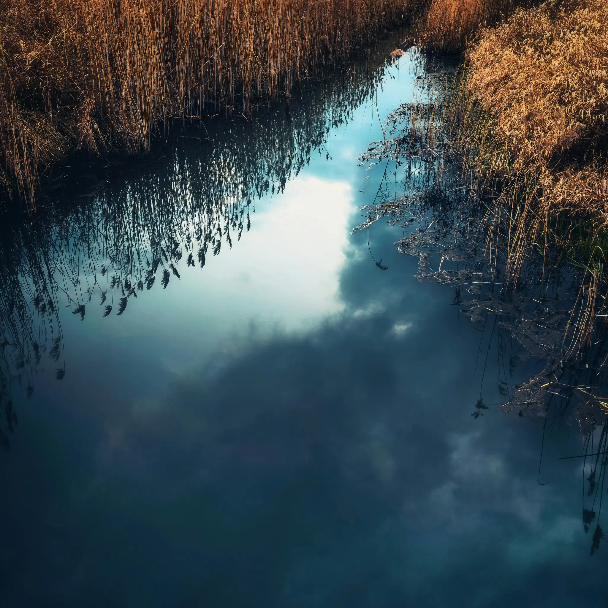 Ein schmaler Kanal aus dunkelblauem Wasser spiegelt einen bewölkten Himmel wider, mit hohen, trockenen, goldbraunen