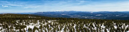 A wide panoramic shot of a snow-covered forest with countless evergreen trees stretching into the distance. Beyond the…