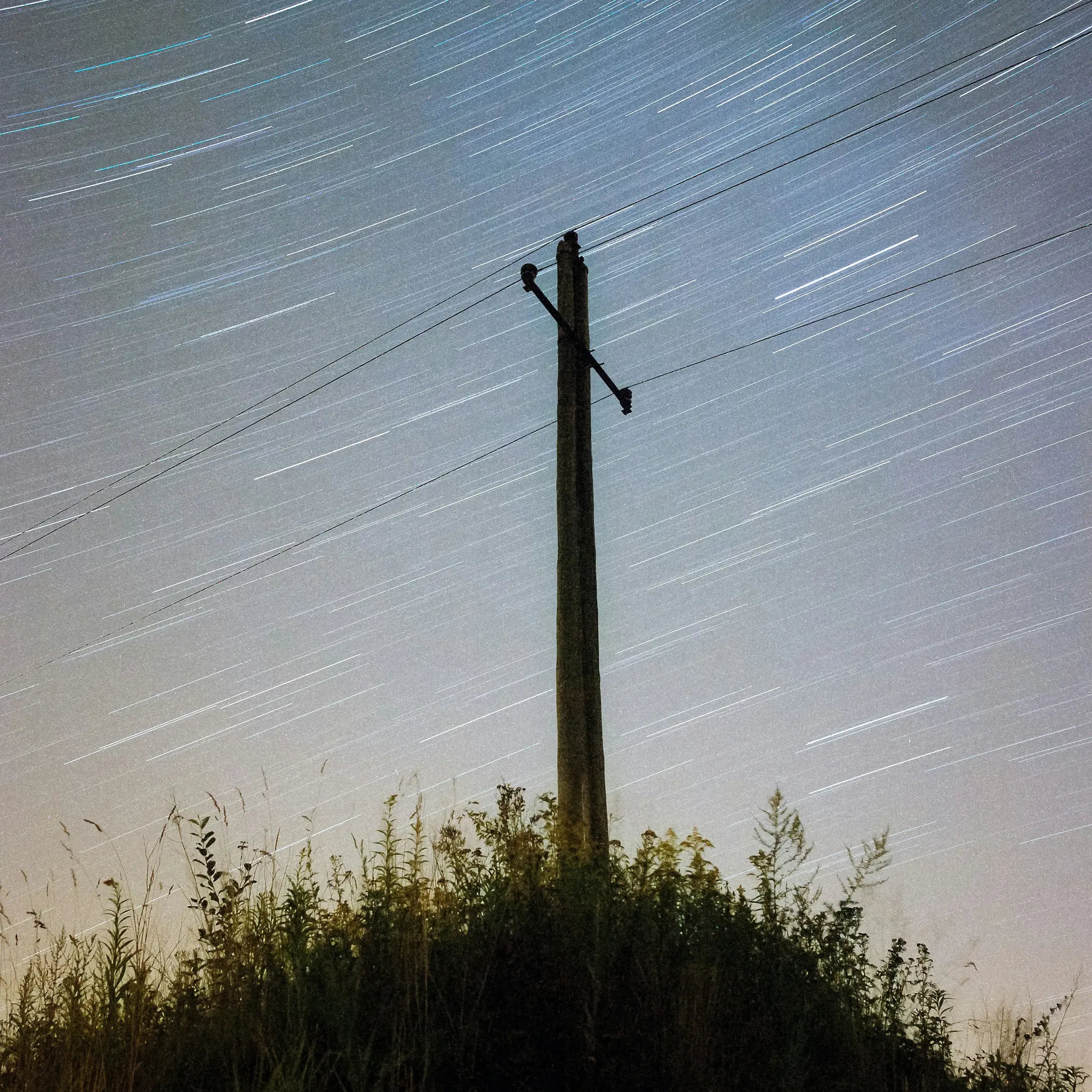 Ein Langzeitbelichtungsfoto zeigt weiße Sternspuren, die sich über einem stilisierten Strommast am dunkelblauen Nachthimmel