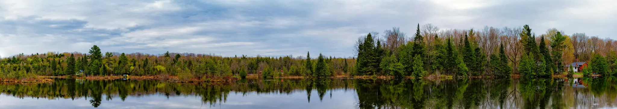 A panoramic photo of a still lake reflecting a dense forest under a cloudy sky. The trees show early spring foliage.